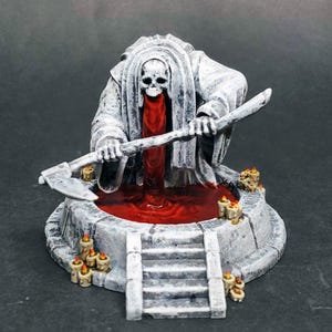 Pode incluir: Uma escultura em miniatura detalhada de uma figura da Morte com rosto de caveira, segurando um machado sobre uma poça de líquido vermelho. A figura está em uma túnica branca, e a cena inclui um poço de pedra com degraus e pequenas velas.