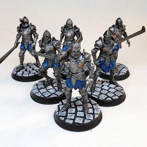 Puede incluir: Un grupo de seis figuras en miniatura de caballeros pintadas de plata que llevan armadura azul y plateada. Las figuras están de pie sobre bases negras con un diseño de adoquines grises.