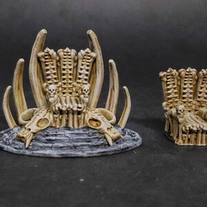 May include: Two miniature bone throne sculptures. The larger throne features a skull-shaped base and curved bone accents. The smaller throne has a similar design. Both are painted in shades of beige and gray.