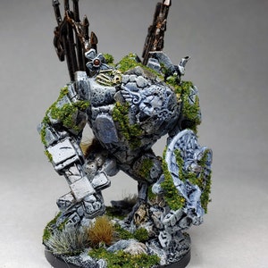 Puede incluir: Una miniatura de golem de piedra gris y blanca con un escudo y una espada. La miniatura está pintada con musgo verde y tiene una textura de piedra detallada.
