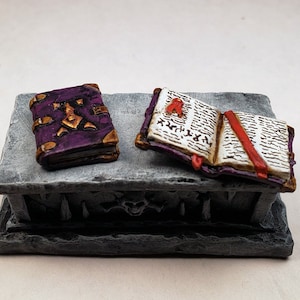 Puede incluir: Dos libros en miniatura sobre una base de piedra gris. Un libro está cerrado y tiene una cubierta morada con detalles dorados. El otro libro está abierto y tiene una cubierta blanca con escritura negra y un marcador de cinta roja.