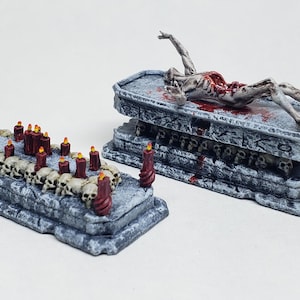 May include: Two miniature, gray, stone-like platforms. One features a row of skulls with red candles, the other has a decaying figure on top. The figure is lying on a stone coffin-like structure, with blood details.