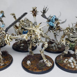 Könnte beinhalten: Eine Sammlung von Miniatur-Skelettfiguren, jede mit einzigartiger Rüstung und Waffen. Einige Skelette haben blaue oder grüne Akzente. Sie stehen auf kleinen, runden Sockeln mit Schädeln und anderen Gelände-Elementen.