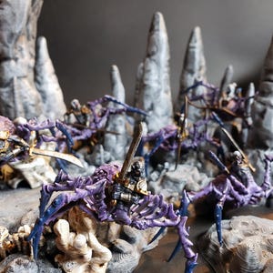 May include: A detailed miniature scene featuring several purple and blue spider-like creatures with swords, set against a backdrop of grey rock formations. The figures are intricately designed, showcasing a fantasy theme.