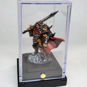 May include: A painted miniature figure of a warrior wearing a red cape and holding a sword, displayed in a clear plastic display case with a black base.