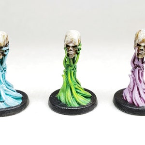 Puede incluir: Tres figuritas de calavera en miniatura con bases de diferentes colores. Las calaveras son blancas con detalles marrones y están unidas a una base azul, verde y morada.