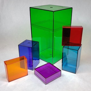 Pode incluir: Uma coleção de caixas de exibição de acrílico transparente e colorido em várias formas e tamanhos. As caixas são em tons de verde, azul, vermelho, laranja e roxo. A caixa maior é uma caixa verde retangular alta. As outras caixas são cubos e prismas.