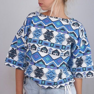 May include: A short-sleeved top with a geometric pattern in shades of blue, teal, and purple on a white background. The top has a wide neckline and a loose fit, suitable for casual wear. The design features repeating shapes.