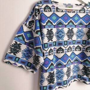 May include: A short-sleeved, white, crew neck shirt with a geometric pattern in shades of blue, teal, and purple. The design features diamond and rectangular shapes. The shirt is hanging on a white hanger.