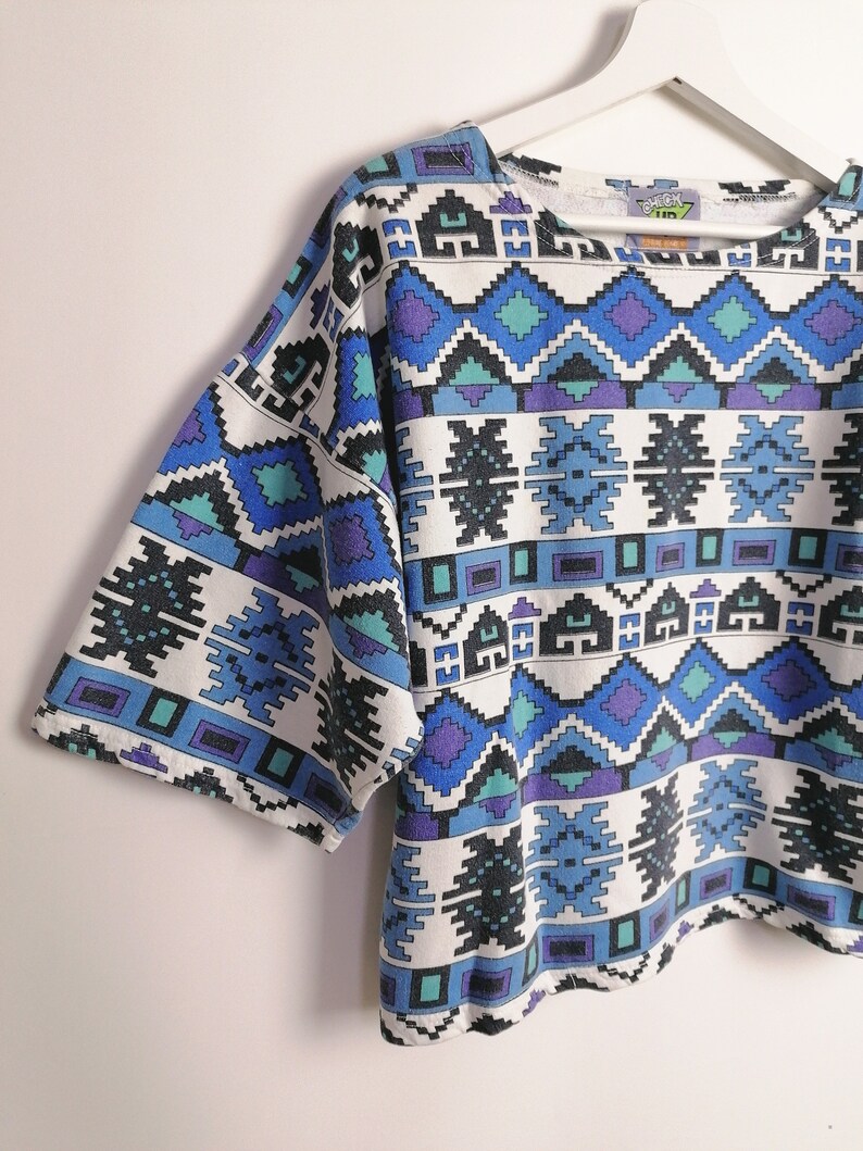 May include: A short-sleeved, white, crew neck shirt with a geometric pattern in shades of blue, teal, and purple. The design features diamond and rectangular shapes. The shirt is hanging on a white hanger.