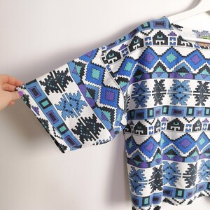 May include: White short-sleeved top with a geometric pattern in shades of blue, teal, and purple. The pattern features diamond and rectangular shapes. The top is hanging on a white hanger.