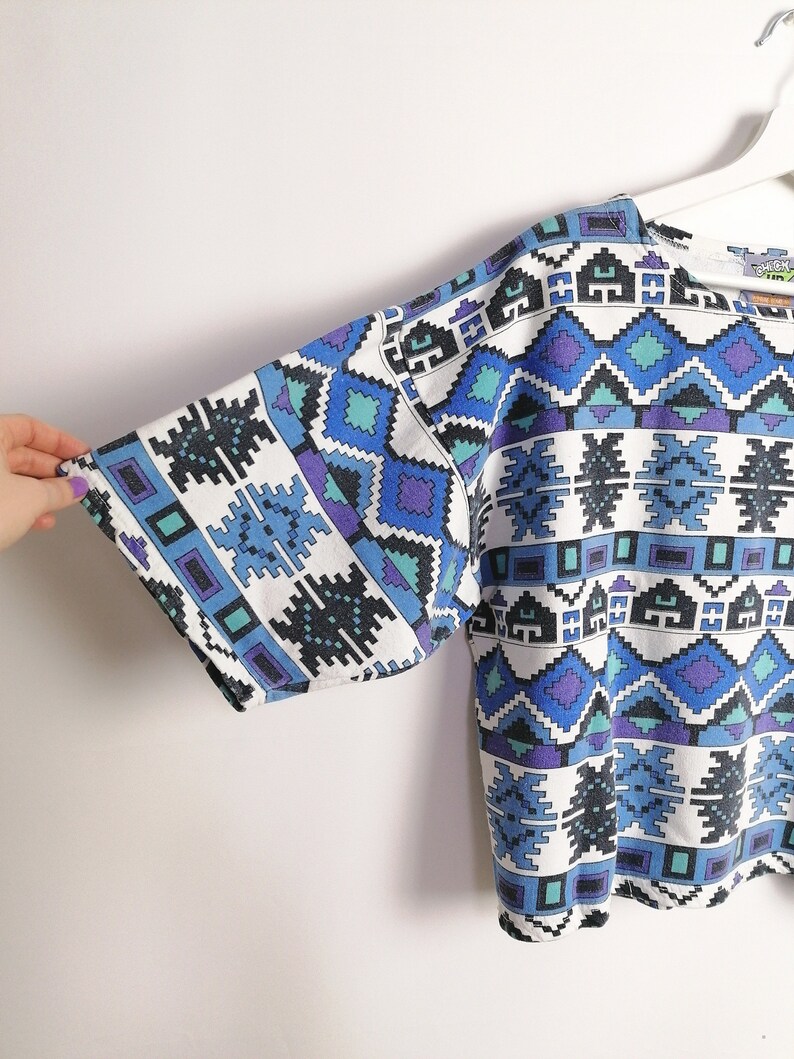 May include: White short-sleeved top with a geometric pattern in shades of blue, teal, and purple. The pattern features diamond and rectangular shapes. The top is hanging on a white hanger.