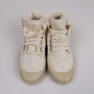 May include: Pair of off-white canvas high-top sneakers. The shoes have a rounded toe, a lace-up closure, and a rubber sole. The canvas material is a light cream color, and the rubber sole is a slightly darker shade.