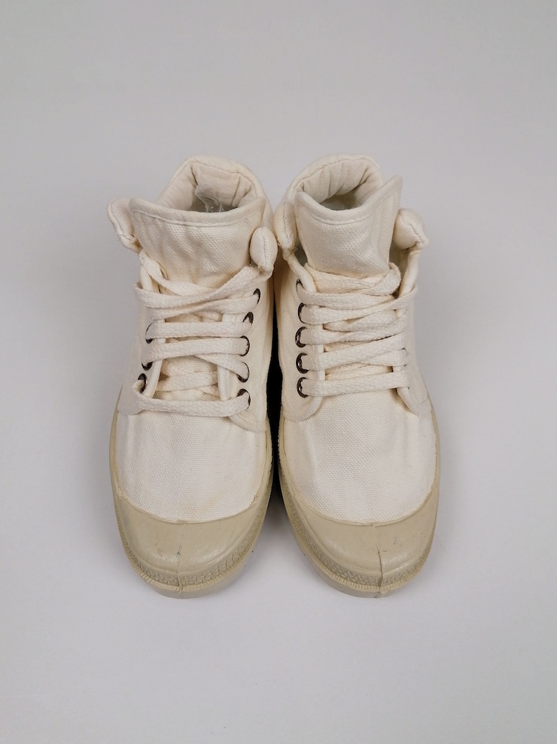 May include: Pair of off-white canvas high-top sneakers. The shoes have a rounded toe, a lace-up closure, and a rubber sole. The canvas material is a light cream color, and the rubber sole is a slightly darker shade.