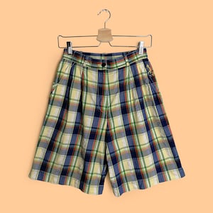 Op de afbeelding: Een geruite short in klassieke stijl, met een geel, blauw, groen en rood geblokt patroon. De short hangt aan een houten hanger tegen een perzikkleurige achtergrond. De short heeft een knoopsluiting en zakken.