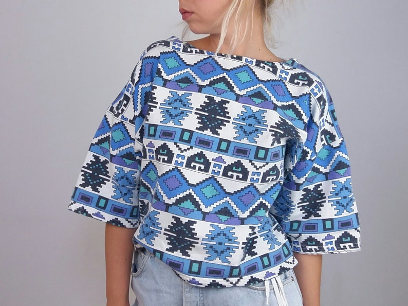 May include: A short-sleeved top with a geometric pattern in shades of blue, teal, and purple on a white background. The top has a wide neckline and a loose fit, suitable for casual wear. The design features repeating shapes.