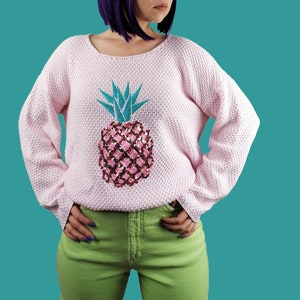 Könnte beinhalten: Hellrosa Pullover mit strukturiertem Strickmuster. Der Pullover hat ein paillettenbesetztes Ananas-Motiv in Rosa, Braun und Türkis. Der Pullover hat eine lockere Passform und lange Ärmel.