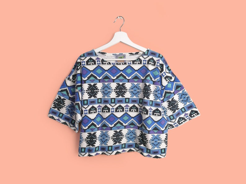 May include: A white, short-sleeved top with a geometric pattern in shades of blue, purple, and black. The top is hanging on a white hanger against a peach background. The pattern includes diamond and rectangular shapes.