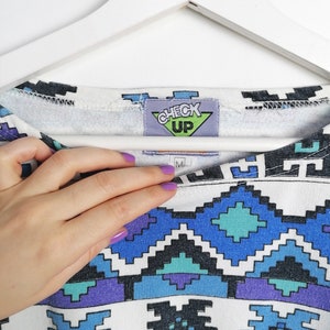 May include: White crew neck sweatshirt with a geometric pattern in shades of blue, teal, purple, and black. The tag reads "CHECK UP". The sweatshirt is hanging on a white hanger.