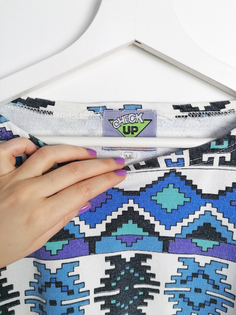 May include: White crew neck sweatshirt with a geometric pattern in shades of blue, teal, purple, and black. The tag reads "CHECK UP". The sweatshirt is hanging on a white hanger.