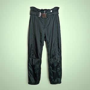 Może przedstawiać: Czarne spodnie narciarskie z wysokim stanem i paskiem z metalową klamrą. Spodnie mają zapięcie na zamek błyskawiczny i pionowe szwy z przodu i po bokach. Spodnie mają lekko błyszczący wygląd i są wykonane z materiału syntetycznego.