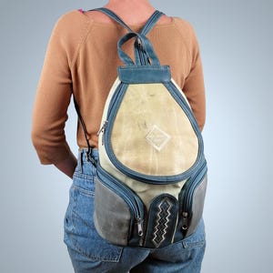 Könnte beinhalten: Ein Vintage-Rucksack mit einzigartigem Design. Die Tasche hat eine cremefarbene Mittelbahn, blaue Zierleisten und graue Seitentaschen. Der Rucksack hat einen rautenförmigen Aufnäher und zwei Reißverschlussfächer.