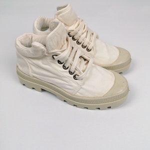 May include: A pair of off-white canvas high-top sneakers. The sneakers have a rounded toe, a lace-up closure with metal eyelets, and a thick, textured rubber sole. The collar is folded down. The overall design is casual and practical.
