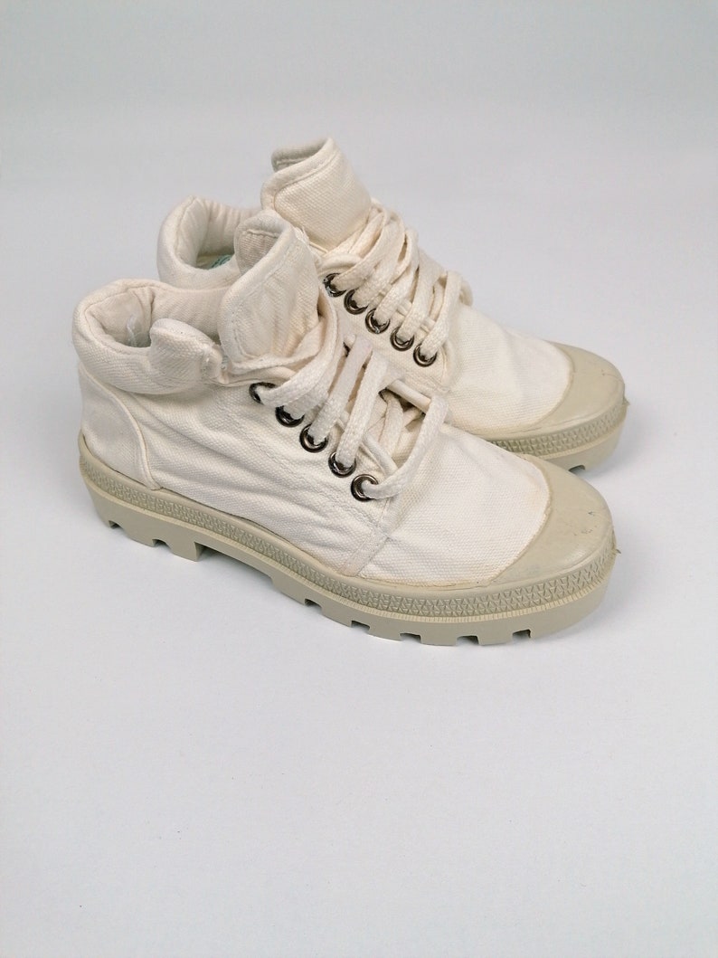May include: A pair of off-white canvas high-top sneakers. The sneakers have a rounded toe, a lace-up closure with metal eyelets, and a thick, textured rubber sole. The collar is folded down. The overall design is casual and practical.