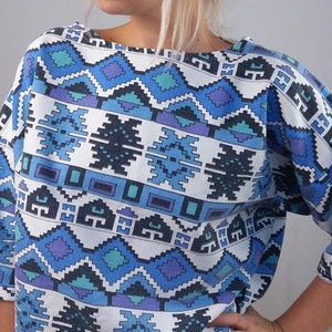 May include: A short-sleeved top with a geometric pattern in shades of blue, purple, and white. The design features repeating diamond and rectangular shapes. The top has a wide neckline and a relaxed fit.