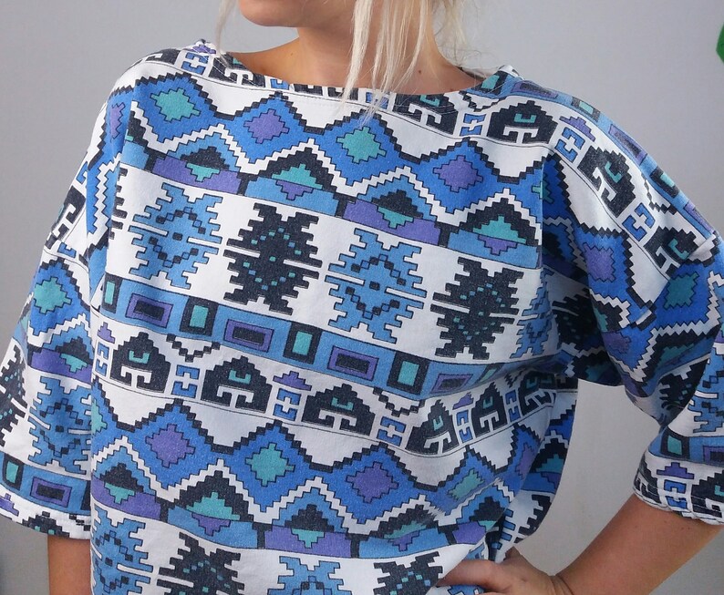May include: A short-sleeved top with a geometric pattern in shades of blue, purple, and white. The design features repeating diamond and rectangular shapes. The top has a wide neckline and a relaxed fit.