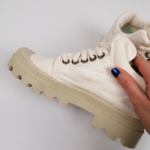 May include: Off-white canvas boot with a thick, beige rubber sole. The boot features metal eyelets for the laces and a padded collar. The sole has a textured, lugged design. The boot is a casual style, suitable for various activities.