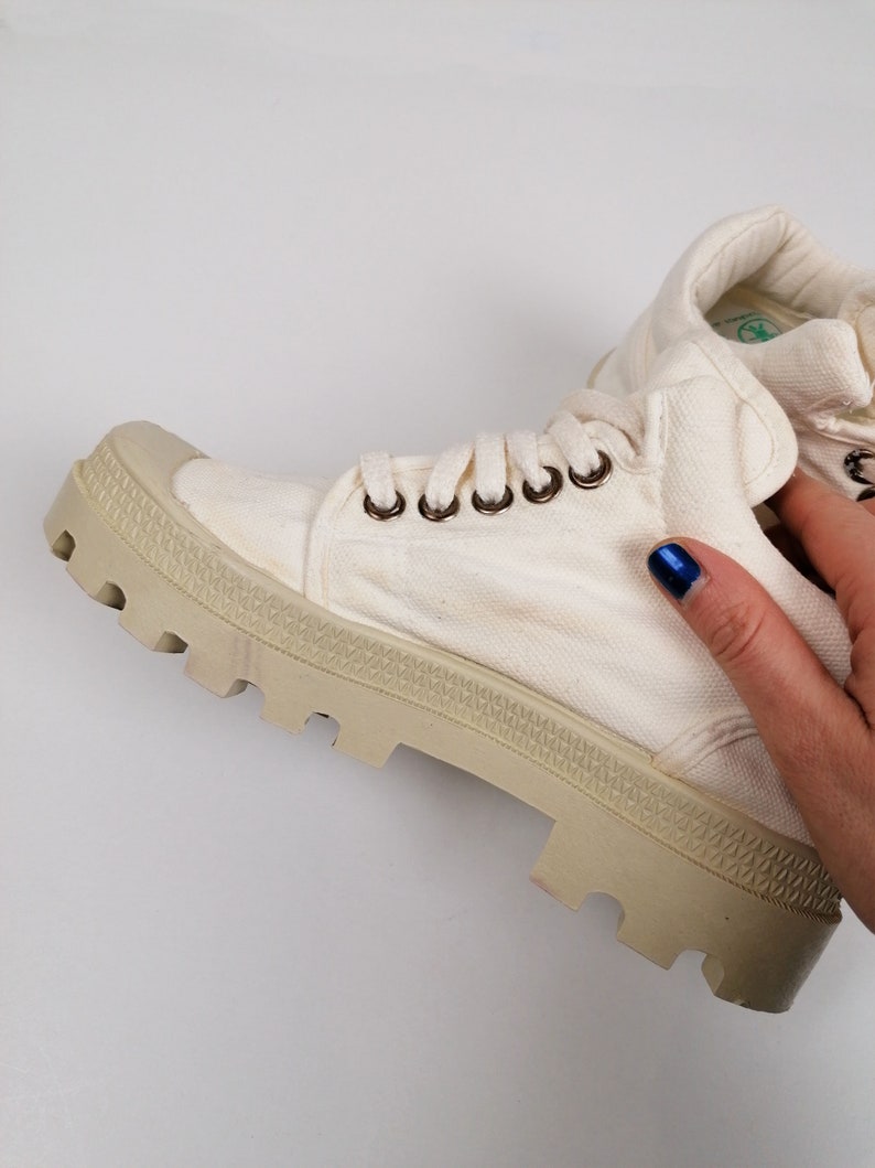 May include: Off-white canvas boot with a thick, beige rubber sole. The boot features metal eyelets for the laces and a padded collar. The sole has a textured, lugged design. The boot is a casual style, suitable for various activities.