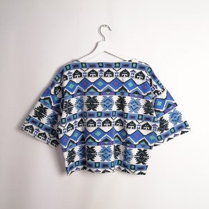 May include: A short-sleeved top with a geometric pattern in shades of blue, teal, and black on a white background. The top is hanging on a white hanger against a white wall. The design features repeating diamond and rectangular shapes.
