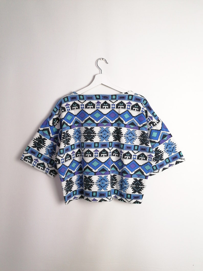 May include: A short-sleeved top with a geometric pattern in shades of blue, teal, and black on a white background. The top is hanging on a white hanger against a white wall. The design features repeating diamond and rectangular shapes.