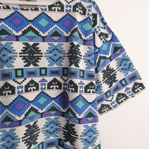May include: A short-sleeved, white t-shirt with a geometric pattern in shades of blue, teal, purple, and black. The pattern features diamond and rectangular shapes. The text "CHECK UP" is visible on the neckline.