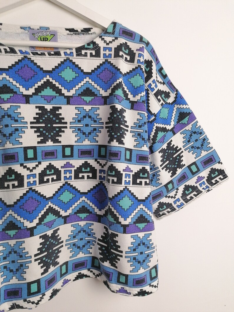 May include: A short-sleeved, white t-shirt with a geometric pattern in shades of blue, teal, purple, and black. The pattern features diamond and rectangular shapes. The text "CHECK UP" is visible on the neckline.