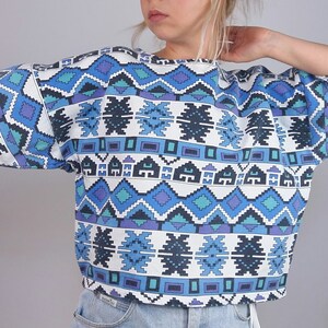 May include: A short-sleeved, cropped top with a geometric pattern in shades of blue, purple, and white. The design features repeating shapes and lines, creating a bold, eye-catching look. The top is made of a soft material.