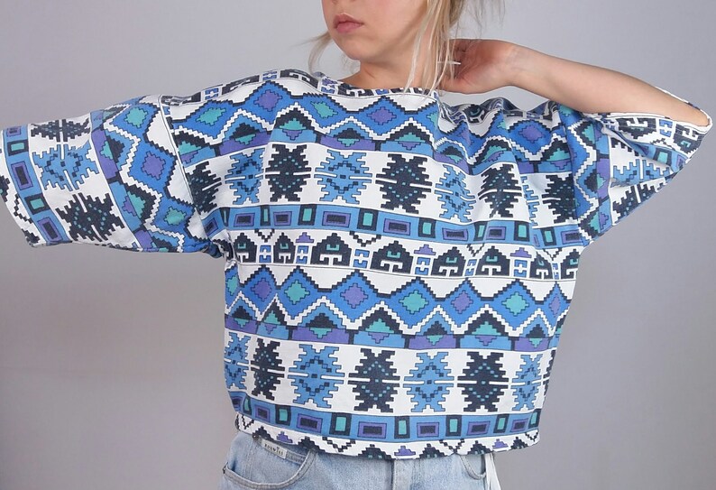May include: A short-sleeved, cropped top with a geometric pattern in shades of blue, purple, and white. The design features repeating shapes and lines, creating a bold, eye-catching look. The top is made of a soft material.