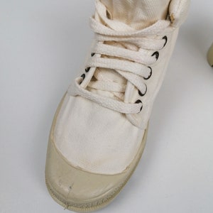 May include: Off-white canvas high-top sneaker with a rubber toe cap and black eyelets. The shoe features white laces and a padded collar. The sneaker is designed for casual wear and has a classic, vintage aesthetic.