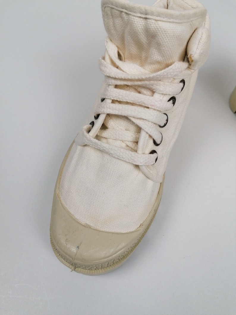 May include: Off-white canvas high-top sneaker with a rubber toe cap and black eyelets. The shoe features white laces and a padded collar. The sneaker is designed for casual wear and has a classic, vintage aesthetic.