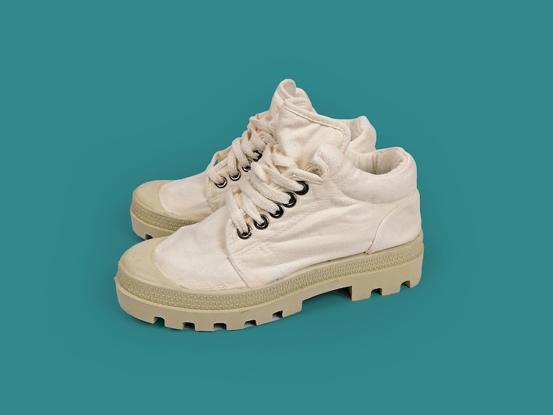 May include: A pair of off-white canvas boots with a chunky, light beige sole. The boots have a lace-up design with black eyelets and a padded collar. The background is a solid teal color.