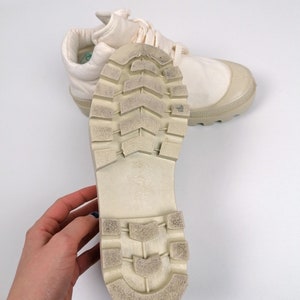 May include: Off-white canvas sneakers with a rubber toe cap and sole. The sole is textured with a tread pattern. The laces are off-white. The sneakers are shown on a white background.