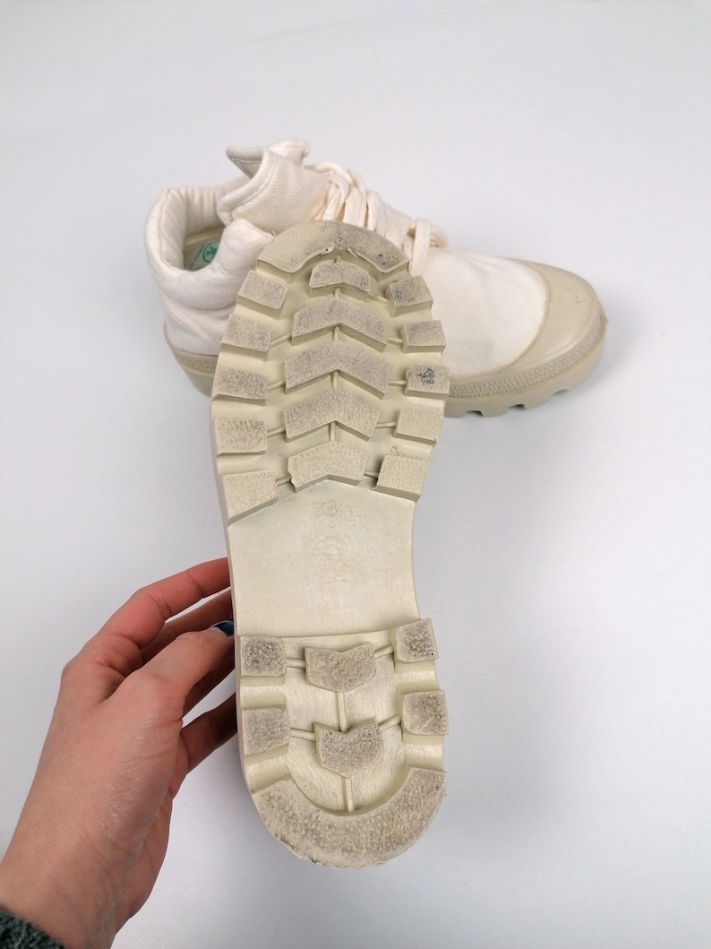 May include: Off-white canvas sneakers with a rubber toe cap and sole. The sole is textured with a tread pattern. The laces are off-white. The sneakers are shown on a white background.