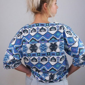May include: A blue, white, and purple patterned top with three-quarter sleeves. The geometric design features diamond and rectangular shapes. The top is paired with light wash denim shorts.