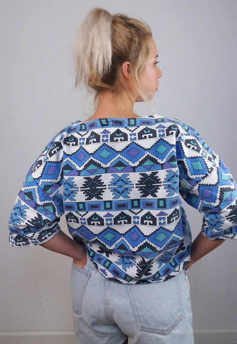 May include: A blue, white, and purple patterned top with three-quarter sleeves. The geometric design features diamond and rectangular shapes. The top is paired with light wash denim shorts.
