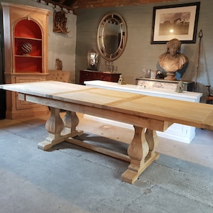 May include: A large, wooden dining table with a natural finish and a rectangular top. The table has two leaves that can be extended to increase its length. The table has a pedestal base with four turned legs.