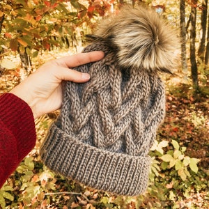 May include: A brown knitted beanie with a fluffy pom pom. The beanie has a cable knit pattern and is photographed in a forest setting with fall foliage.