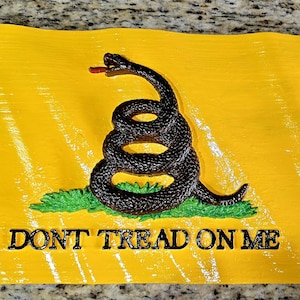 Puede incluir: Un letrero amarillo con una serpiente negra enroscada sobre hierba verde. El texto "DONT TREAD ON ME" está debajo. La serpiente tiene una lengua roja y está centrada en el letrero.