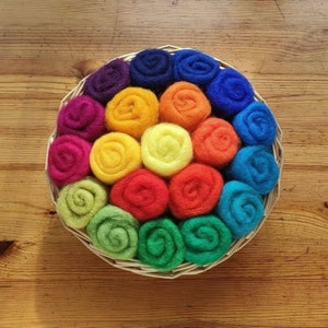 Pode incluir: Um sortimento arco-íris de 15 bolas de lã crua em uma cesta de vime. As bolas são dispostas em círculo, com as cores passando do roxo para o vermelho, laranja, amarelo, verde e azul.