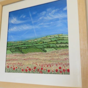 May include: A framed landscape painting depicting a field of red poppies, a wheat field, and green hills under a blue sky with clouds. The artwork is presented in a light wooden frame. The painting evokes a rural scene, with a focus on natural elements.